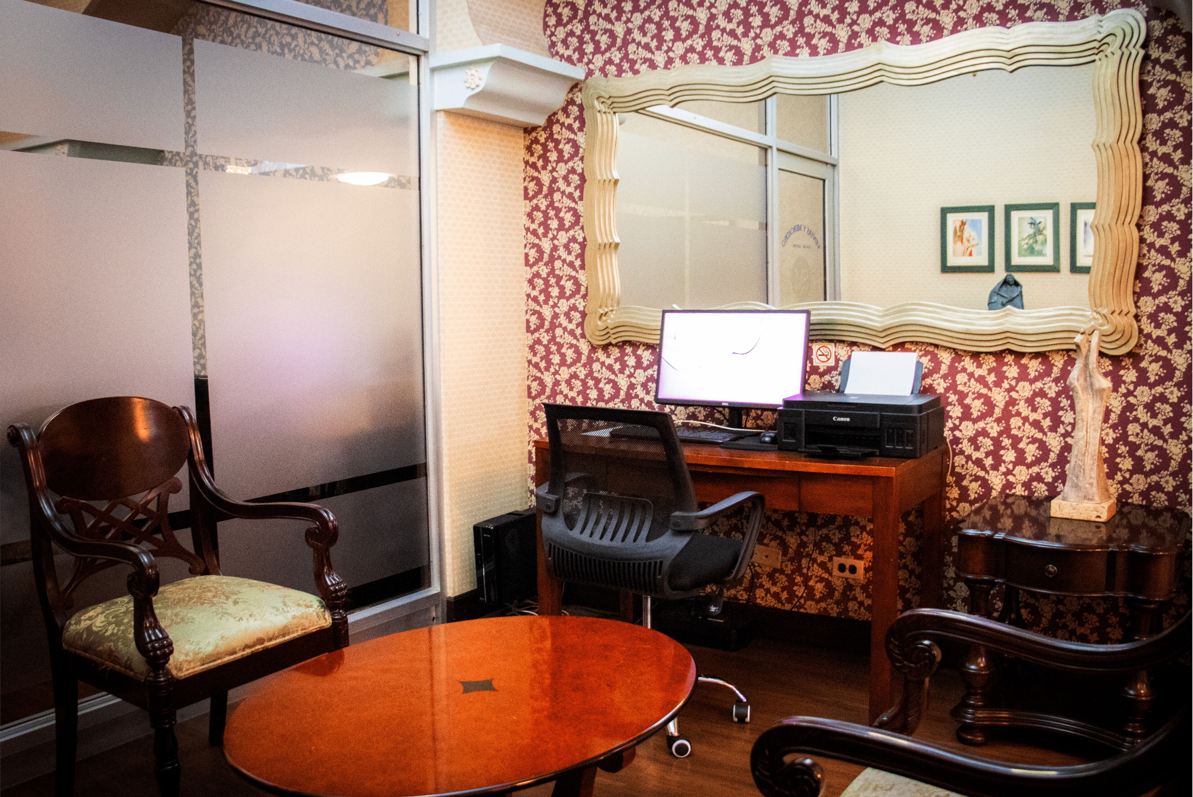 A room with a wooden chair, a desk with a computer, a mirror, and patterned wallpaper.