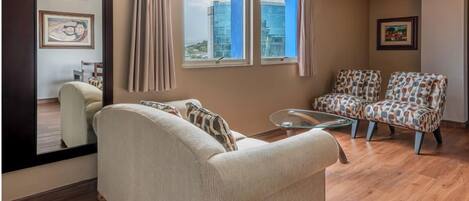 Una sala de estar con un sofá color beige, sillones estampados, una mesa de centro de vidrio y un espejo grande que refleja la habitación.