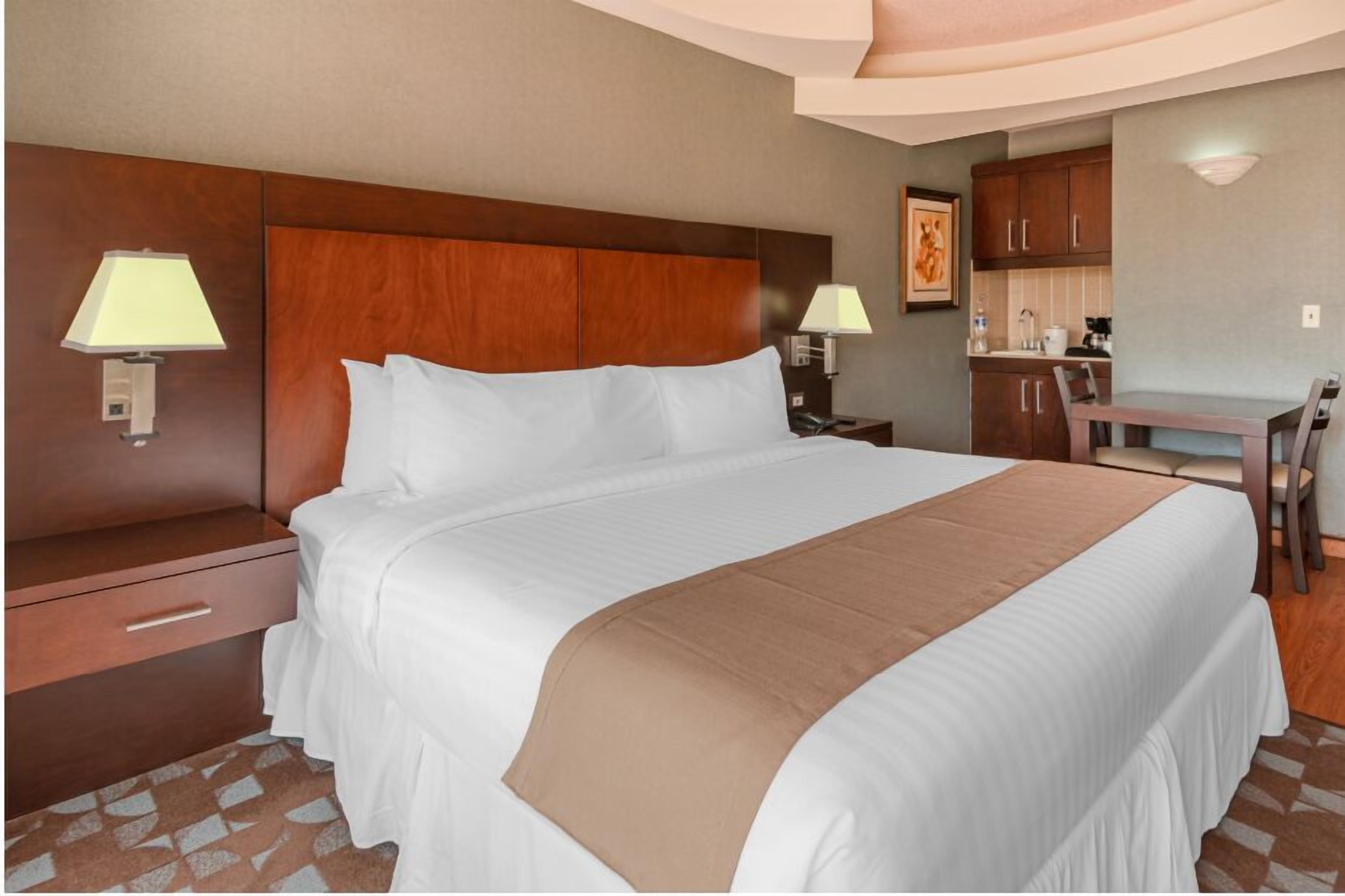 Una habitación de hotel con una cama grande, cabecera de madera, escritorio con silla y kitchenette.