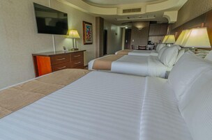 Habitación de hotel con tres camas, un televisor, un escritorio con cajones y una kitchenette al fondo.