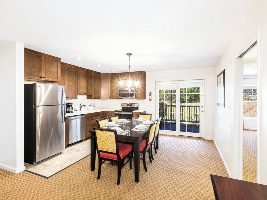 A modern kitchen with a stainless steel refrigerator, wooden cabinets, a dining table set for six, and a view of the backyard through glass doors.