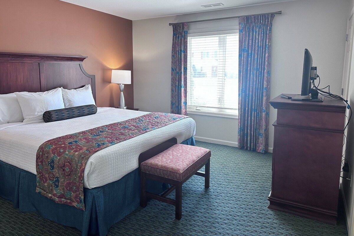 Een hotelkamer met een groot bed, een bureau met een computer, een raam met gordijnen en een lamp.