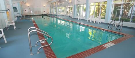 Una piscina interior con agua cristalina, rodeada de ventanas y sillas blancas.