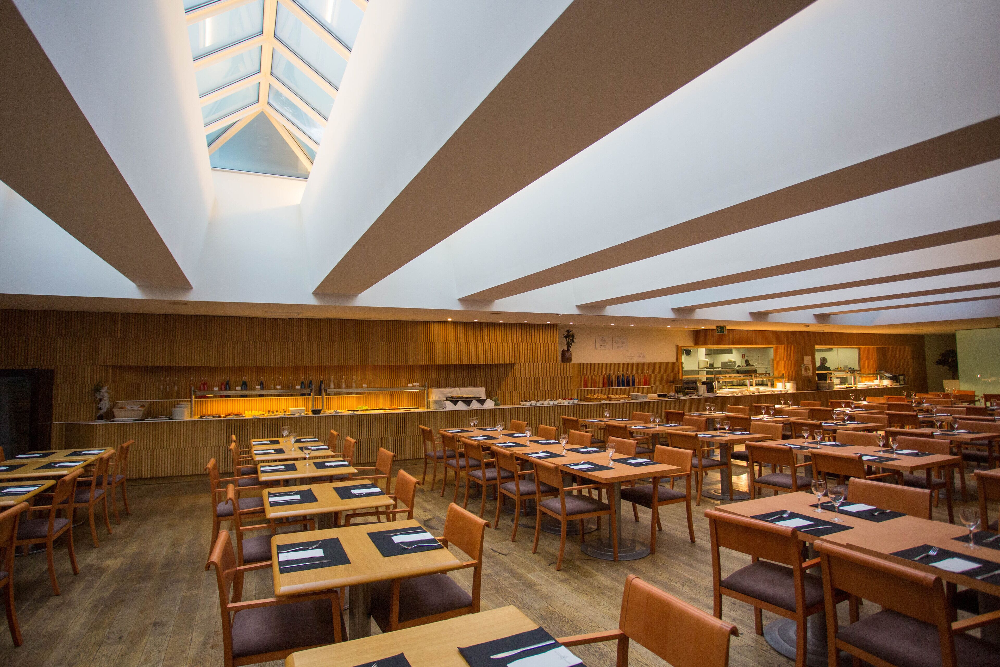 Ruime eetruimte met meerdere tafels en stoelen, een centrale bar en een hoge plafond met dakramen.
