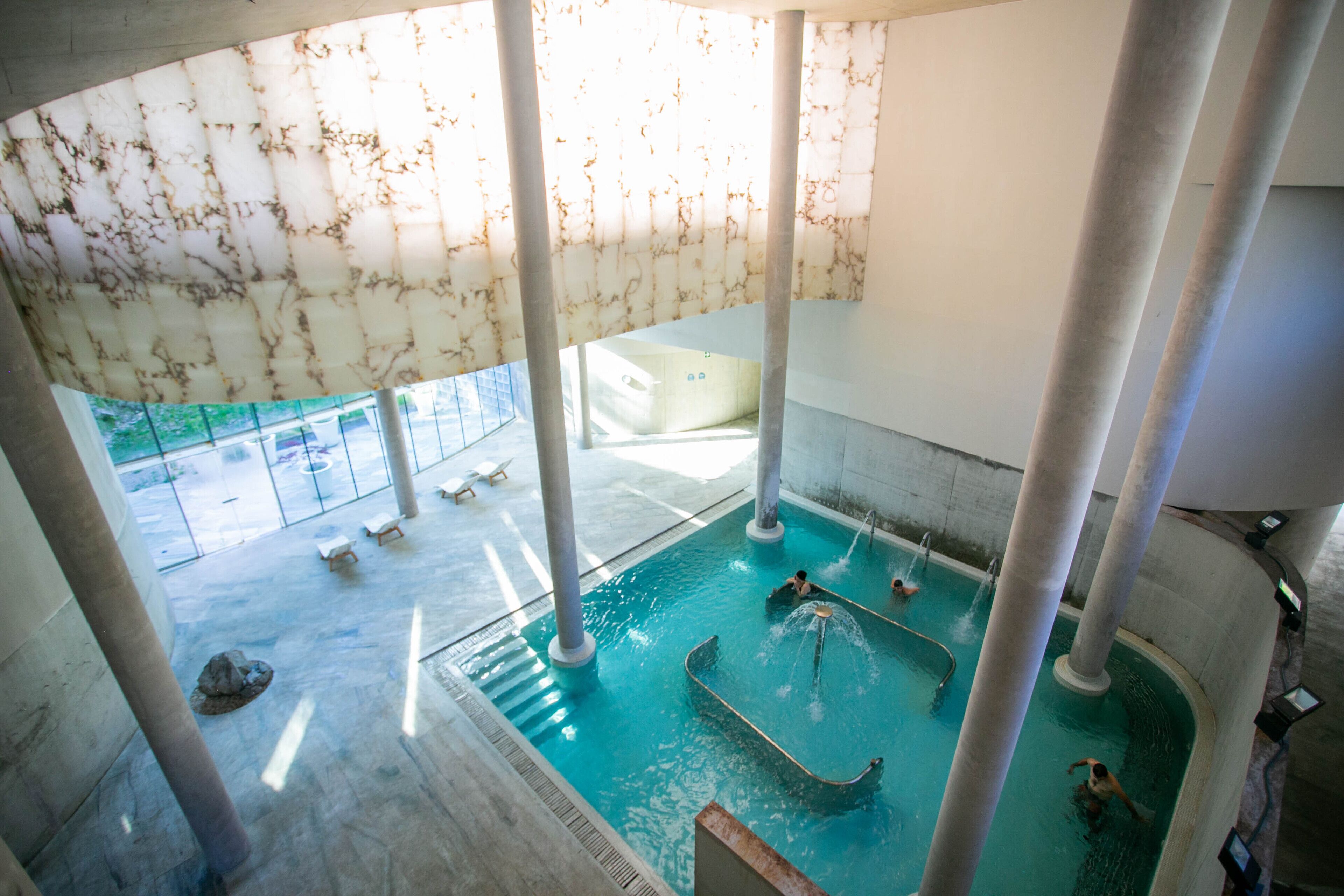 Uma grande piscina interior com água cristalina, rodeada por colunas, e vista para área externa com espreguiçadeiras.
