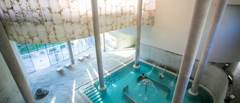 Uma grande piscina interior com água cristalina, rodeada por colunas, e vista para área externa com espreguiçadeiras.
