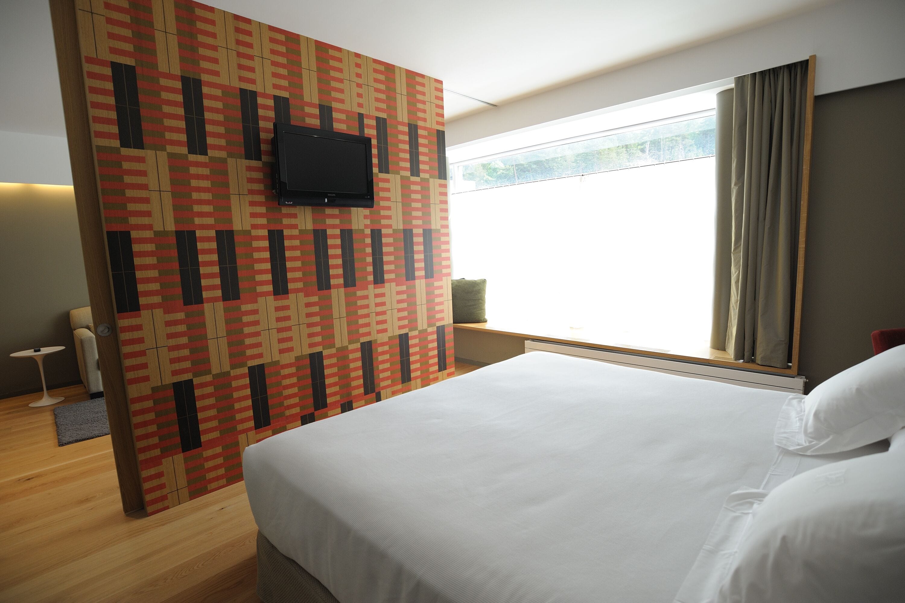 Habitación de hotel con una cama grande, un televisor en una pared estampada, una ventana con cortinas y una mesita con una lámpara.