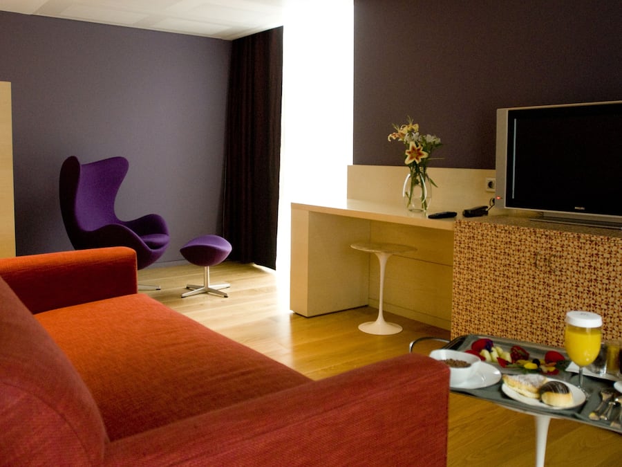Un salon moderne avec un canapé rouge, un fauteuil violet, une petite table ronde sur laquelle sont disposés des éléments pour le petit-déjeuner, et un téléviseur posé sur un meuble.
