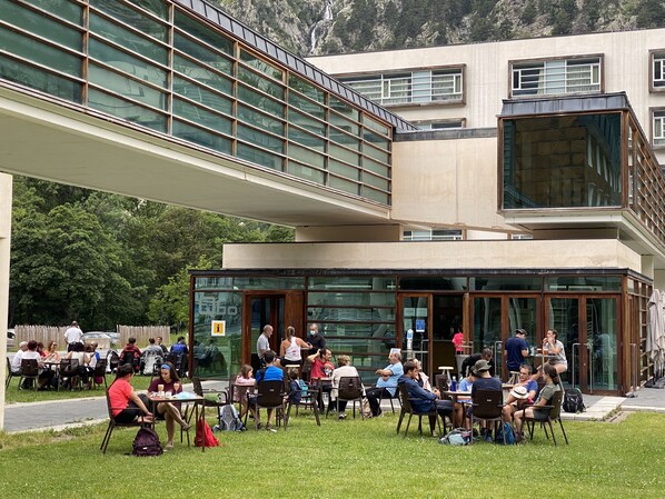 Ein Straßencafé mit Tischen und Stühlen, ein modernes Gebäude mit großen Fenstern und eine Rasenfläche davor.