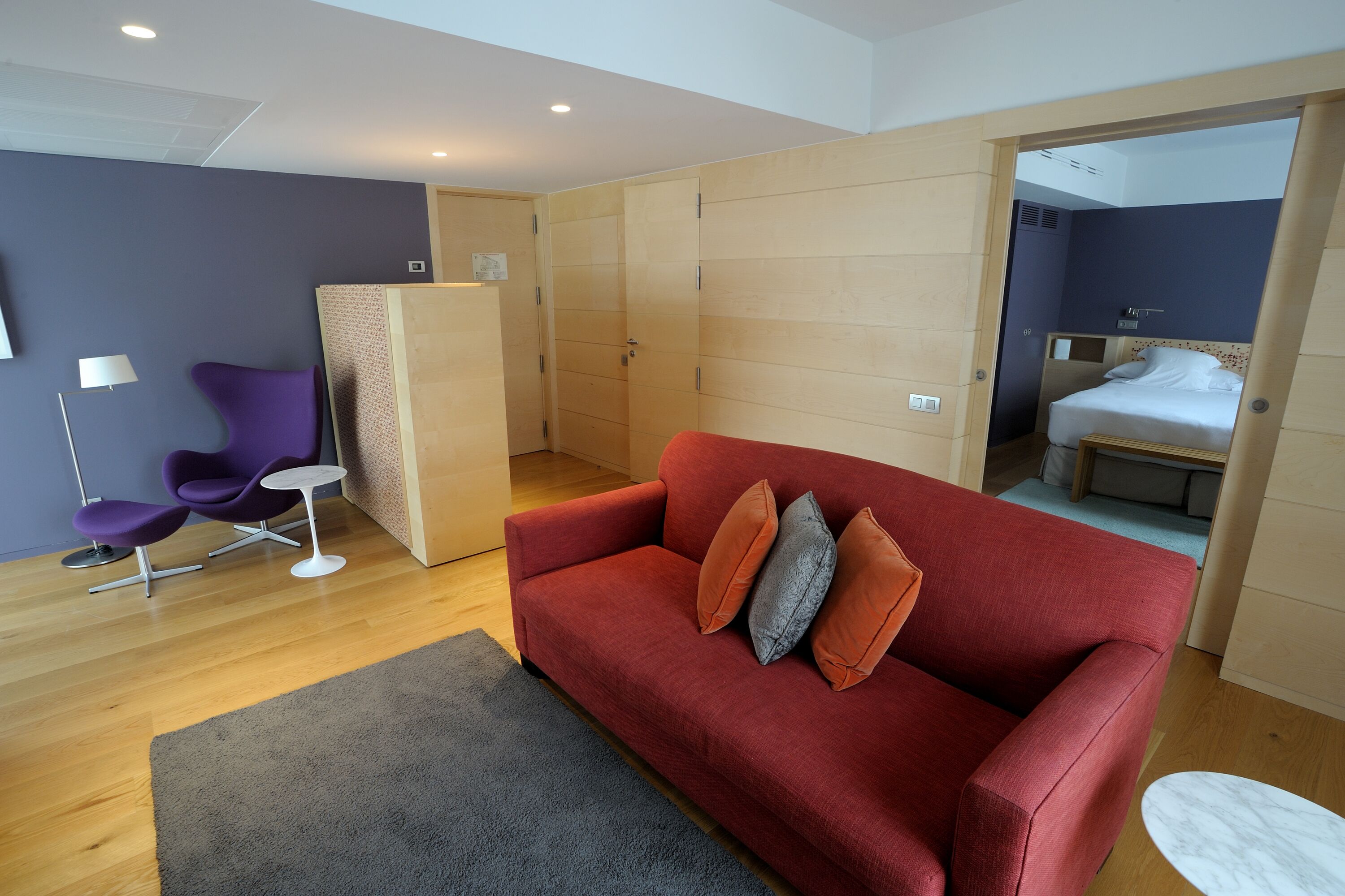 A modern room with a red sofa, a small round table, purple chairs, and a bed in the adjacent room.