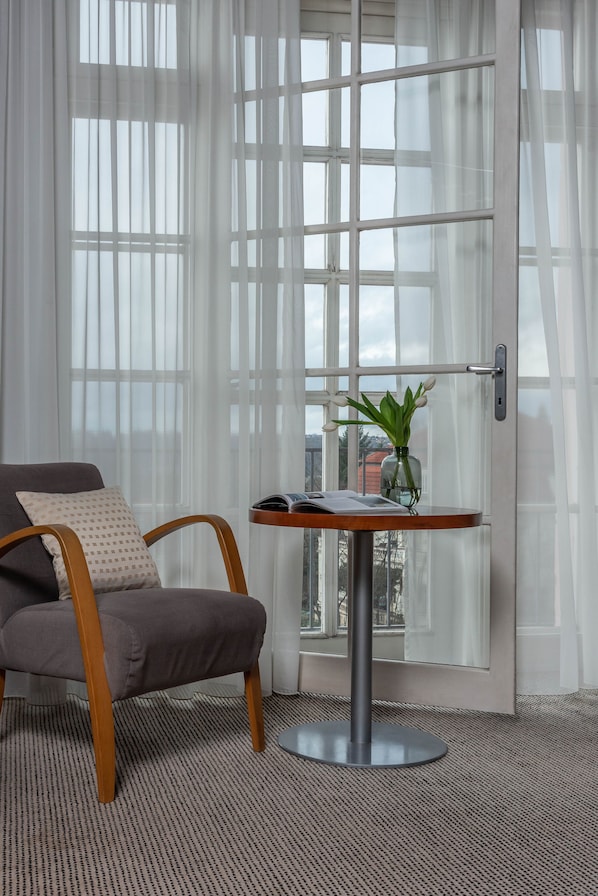 Una habitación con una silla gris, una mesita redonda con un libro y un jarrón de flores, y cortinas de encaje.