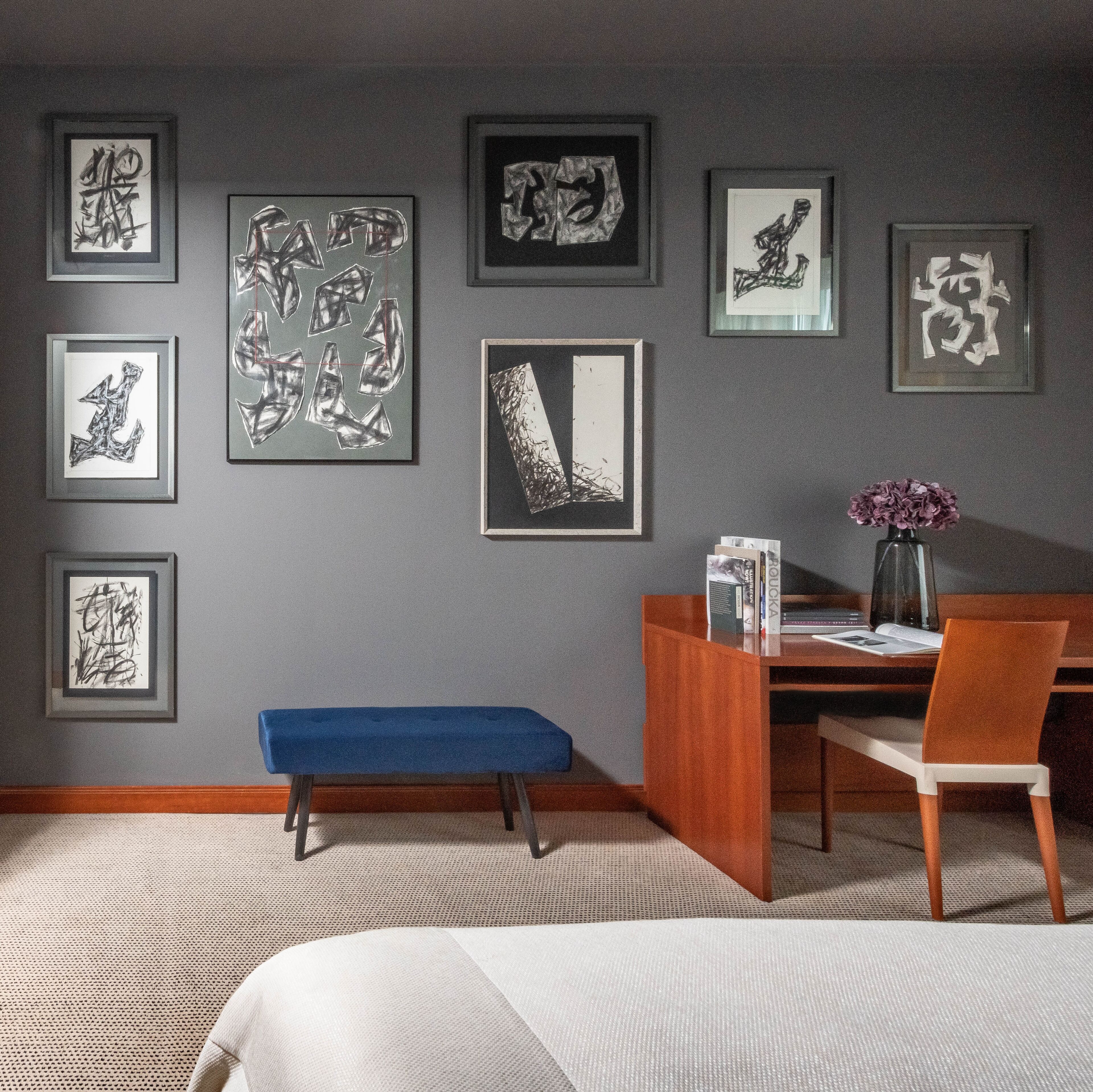 A room with a desk, chair, and bench. There are framed artworks on the wall.