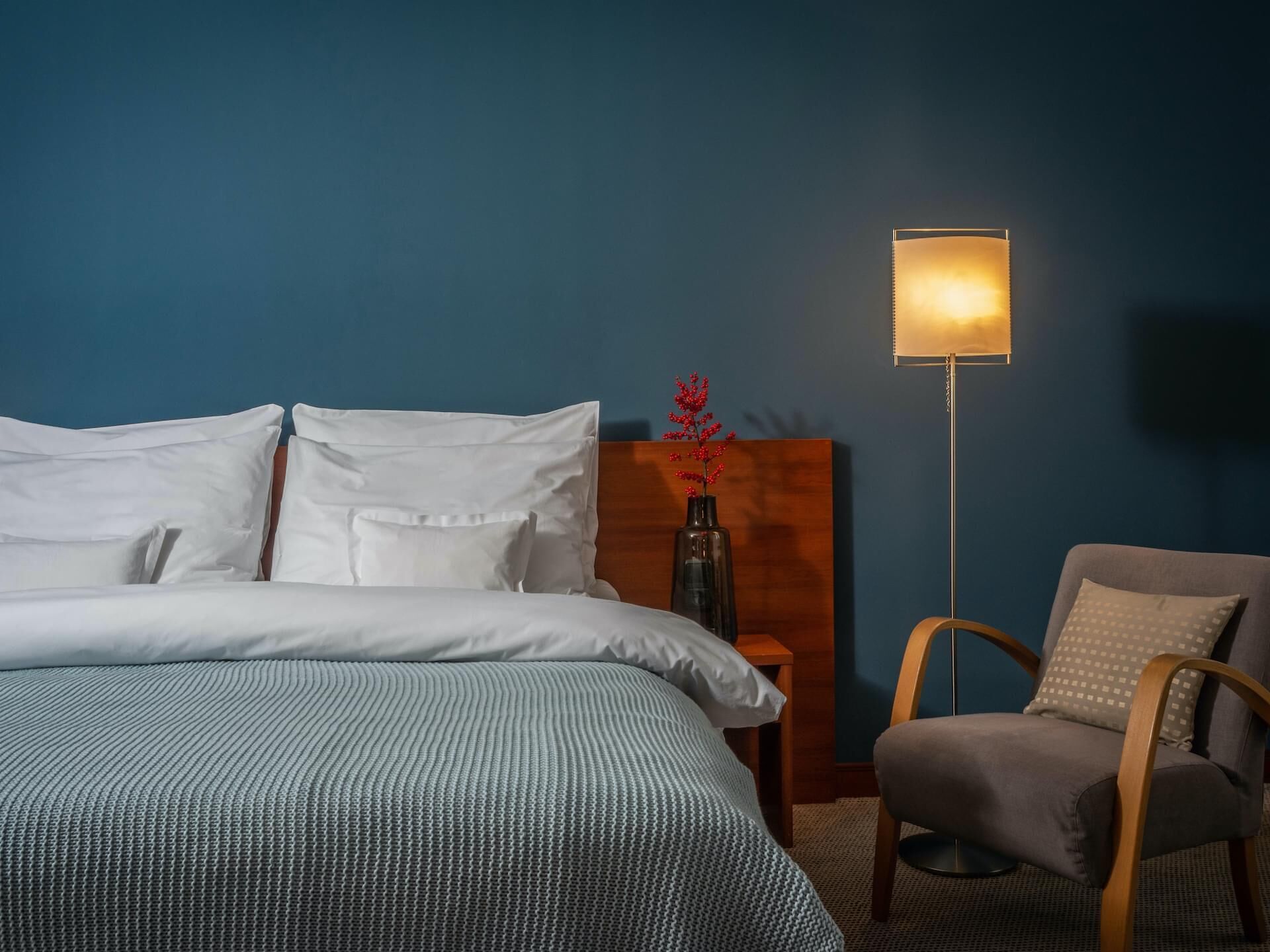 A neatly made bed with white linens, a wooden nightstand with a vase of red flowers, a floor lamp, and a grey armchair in a room with blue walls.