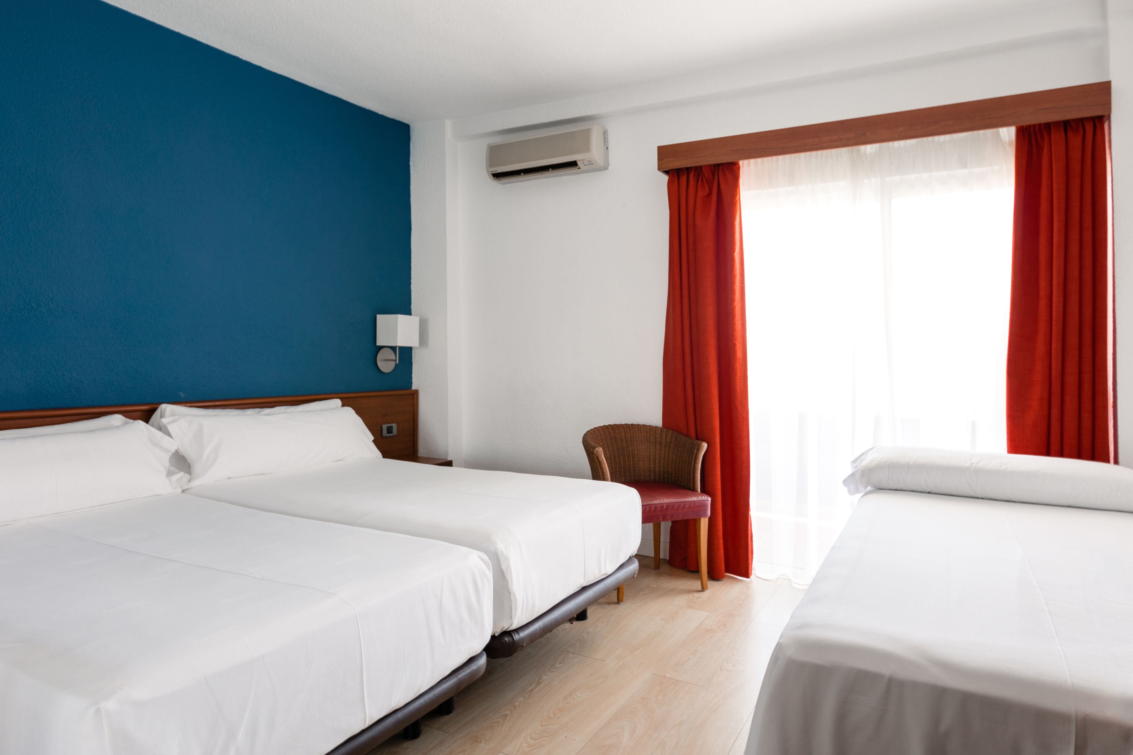 Habitación de hotel con dos camas, una silla, una mesita de noche, un equipo de aire acondicionado y una ventana con cortinas rojas.