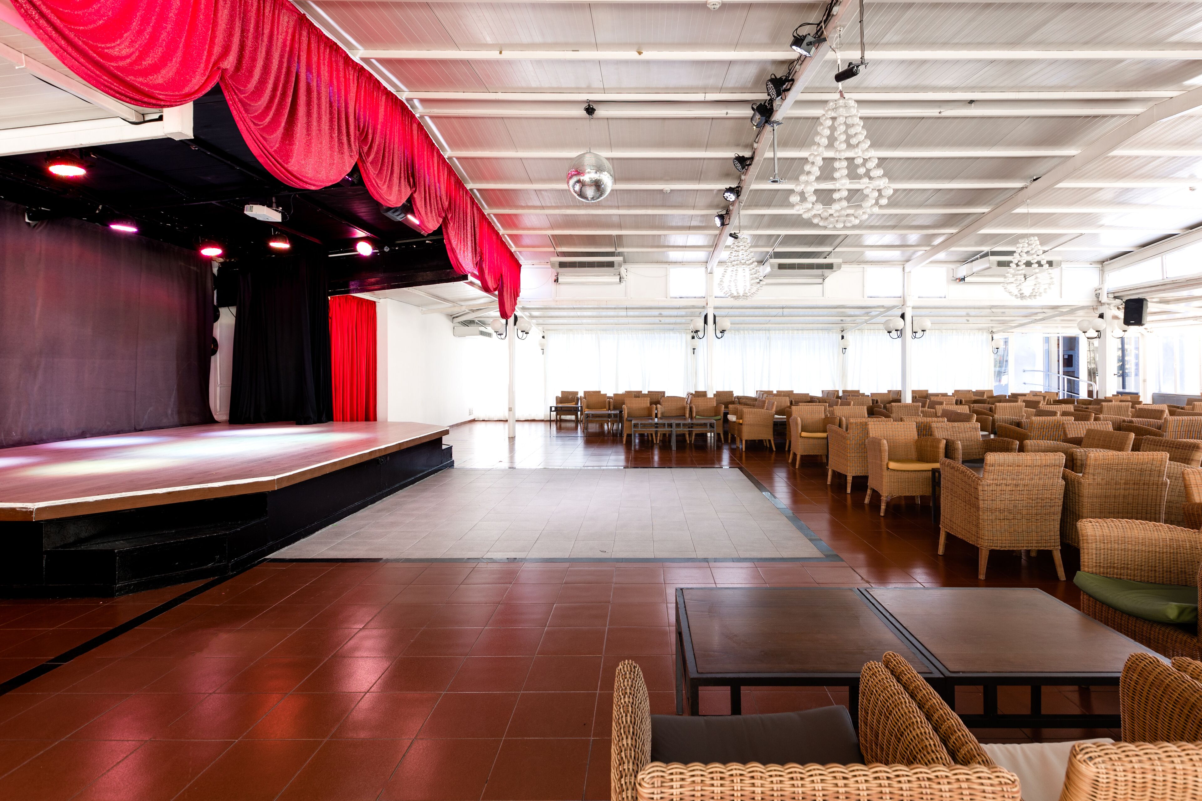 Un salón amplio con un escenario, filas de sillas y una cortina roja.