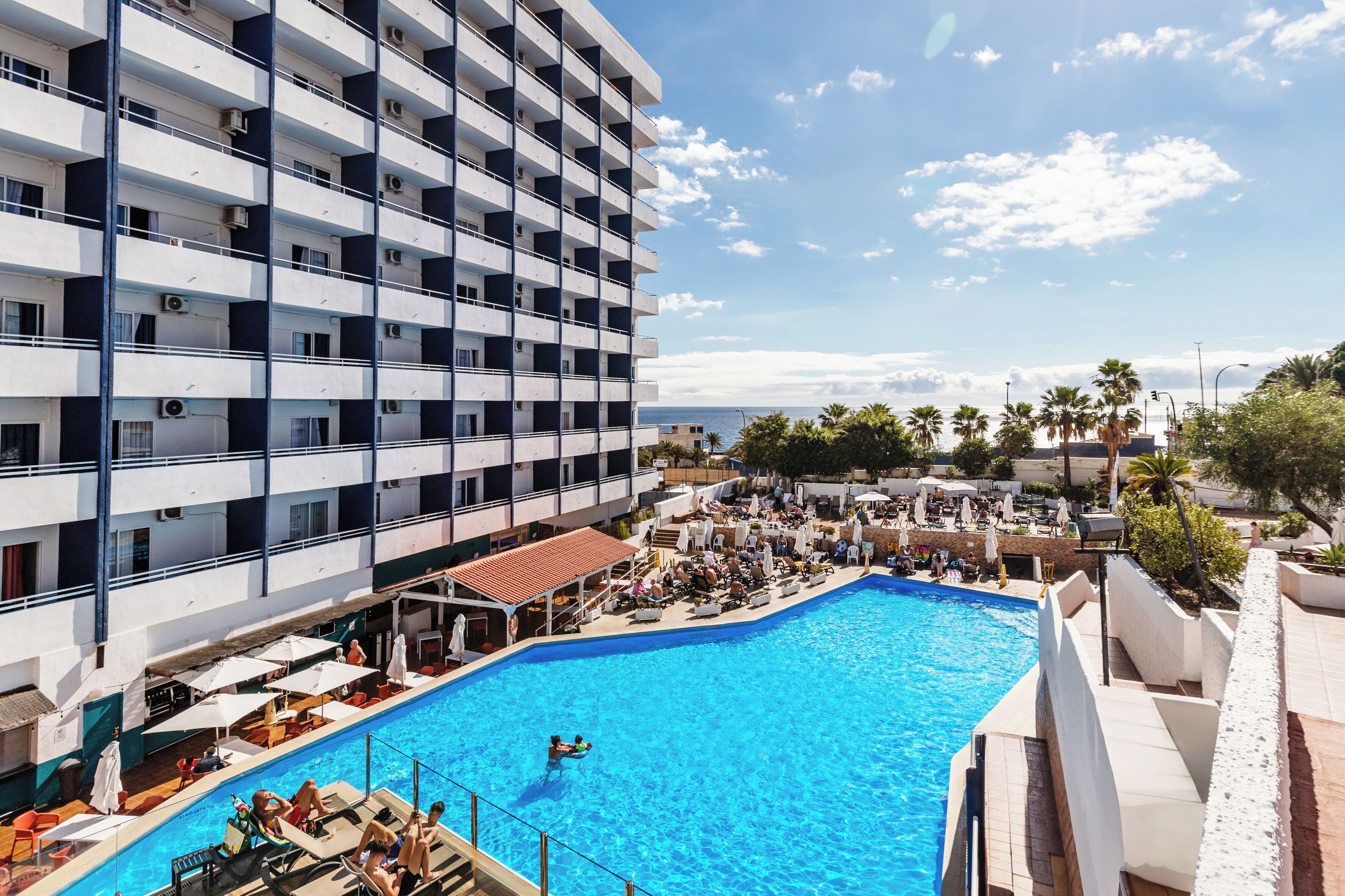 Un hotel con una amplia piscina, varios balcones y vista al mar.