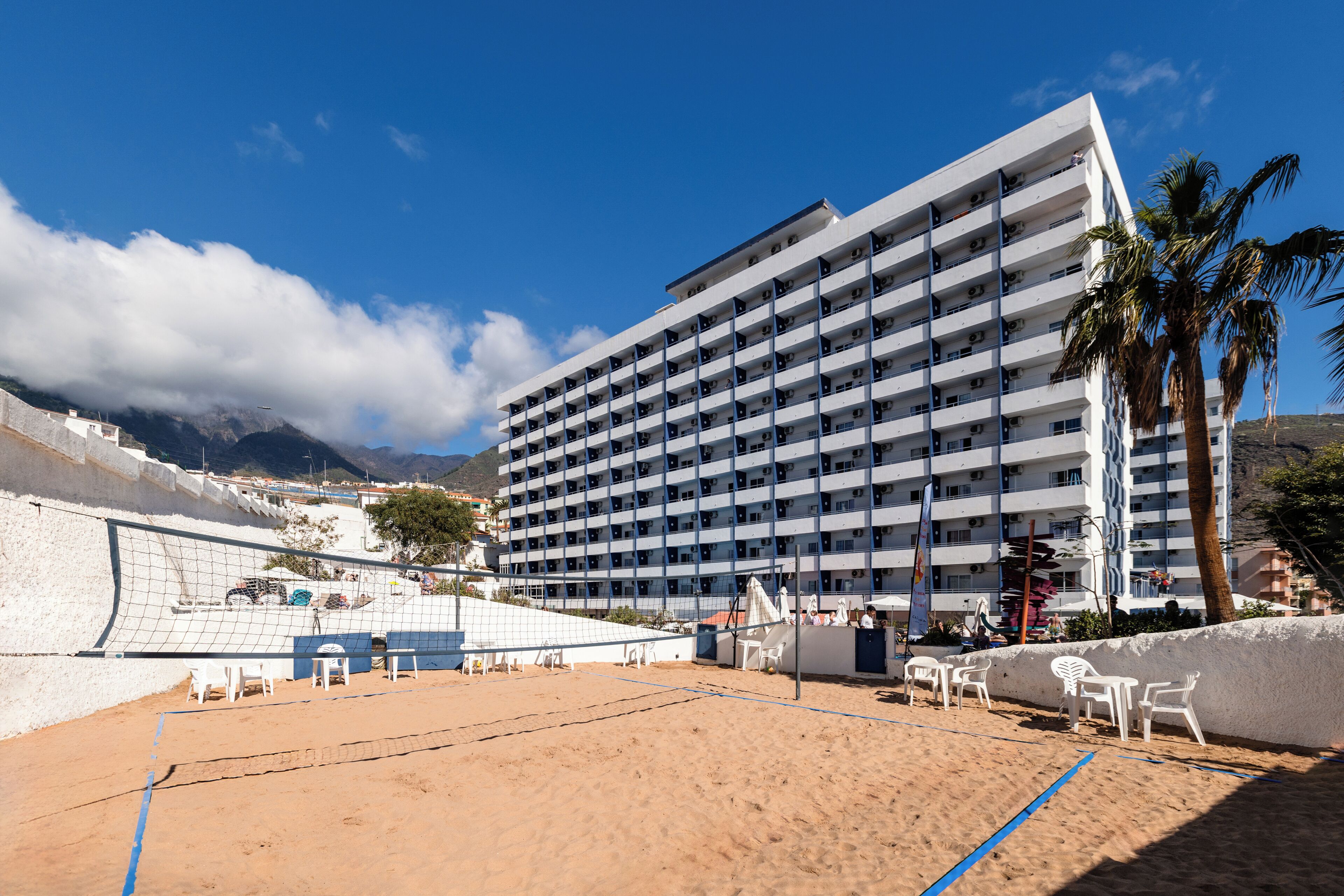 A beach volleyball court with a net, surrounded by white chairs and a building with multiple balconies in the background.