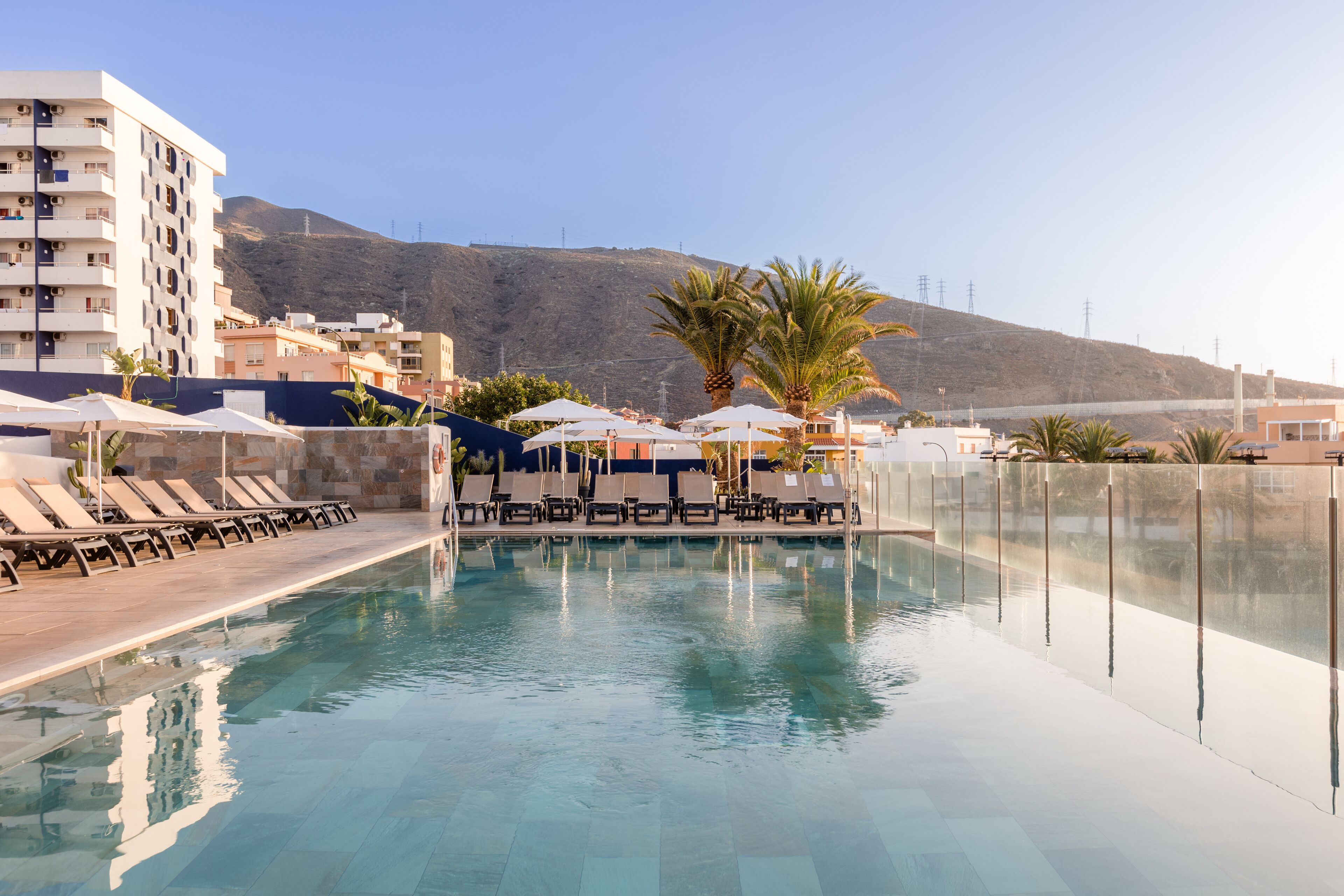 Una piscina con sillas de descanso y sombrillas, rodeada de edificios y palmeras.