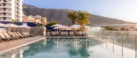 A swimming pool with lounge chairs and umbrellas, surrounded by buildings and palm trees.