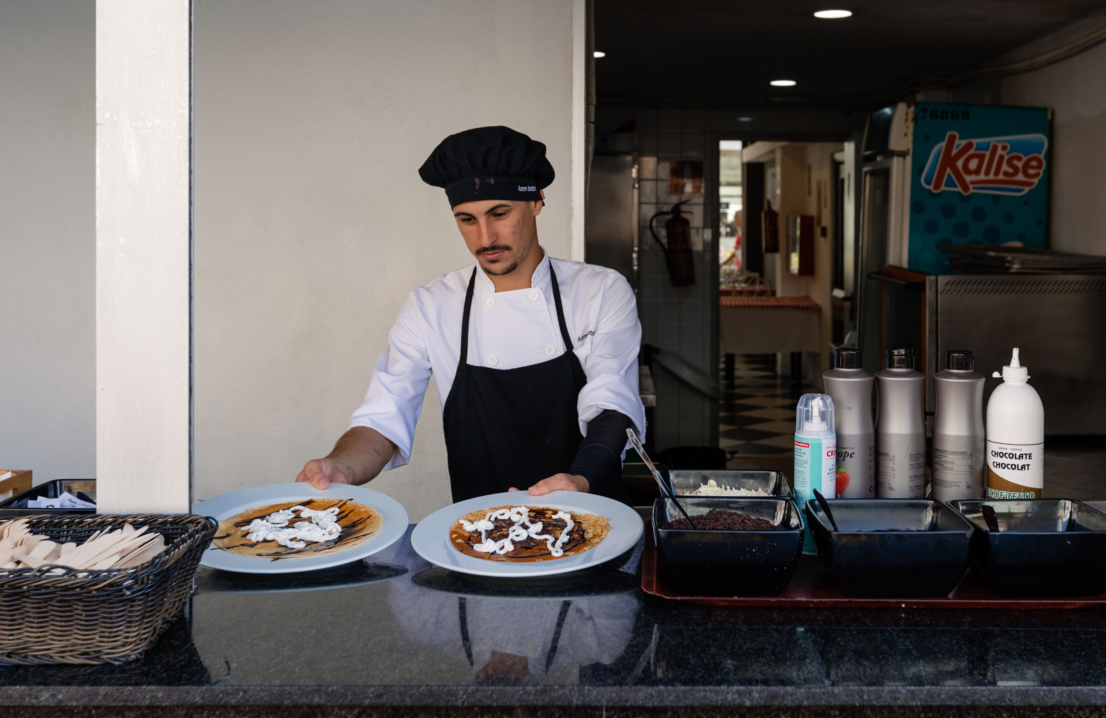 Un chef, vêtu d’une blouse blanche et d’un tablier noir, prépare des plats derrière un comptoir où l’on aperçoit des assiettes remplies de nourriture, un panier d’ustensiles et des condiments.