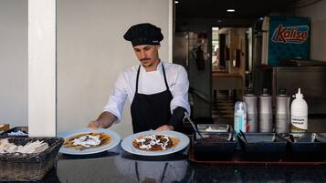 Un chef, vêtu d’une blouse blanche et d’un tablier noir, prépare des plats derrière un comptoir où l’on aperçoit des assiettes remplies de nourriture, un panier d’ustensiles et des condiments.