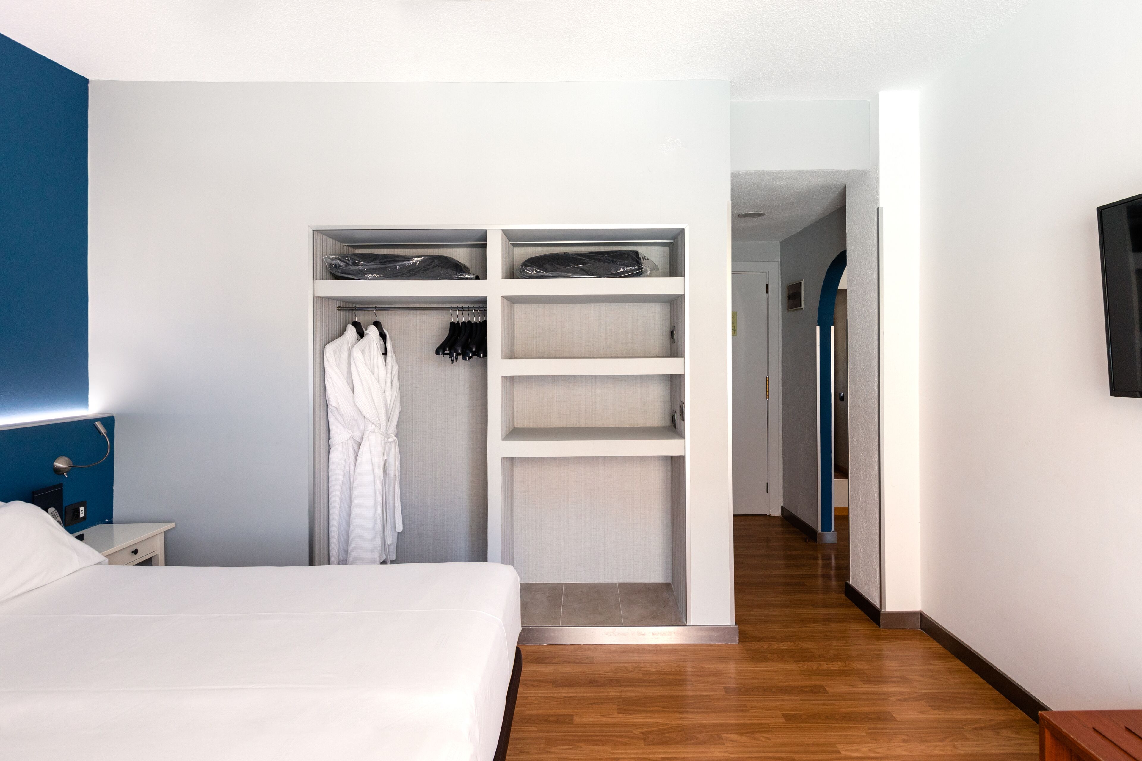 A hotel room with a neatly made bed, a walk-in closet with shelves and hanging space, and a wooden floor.