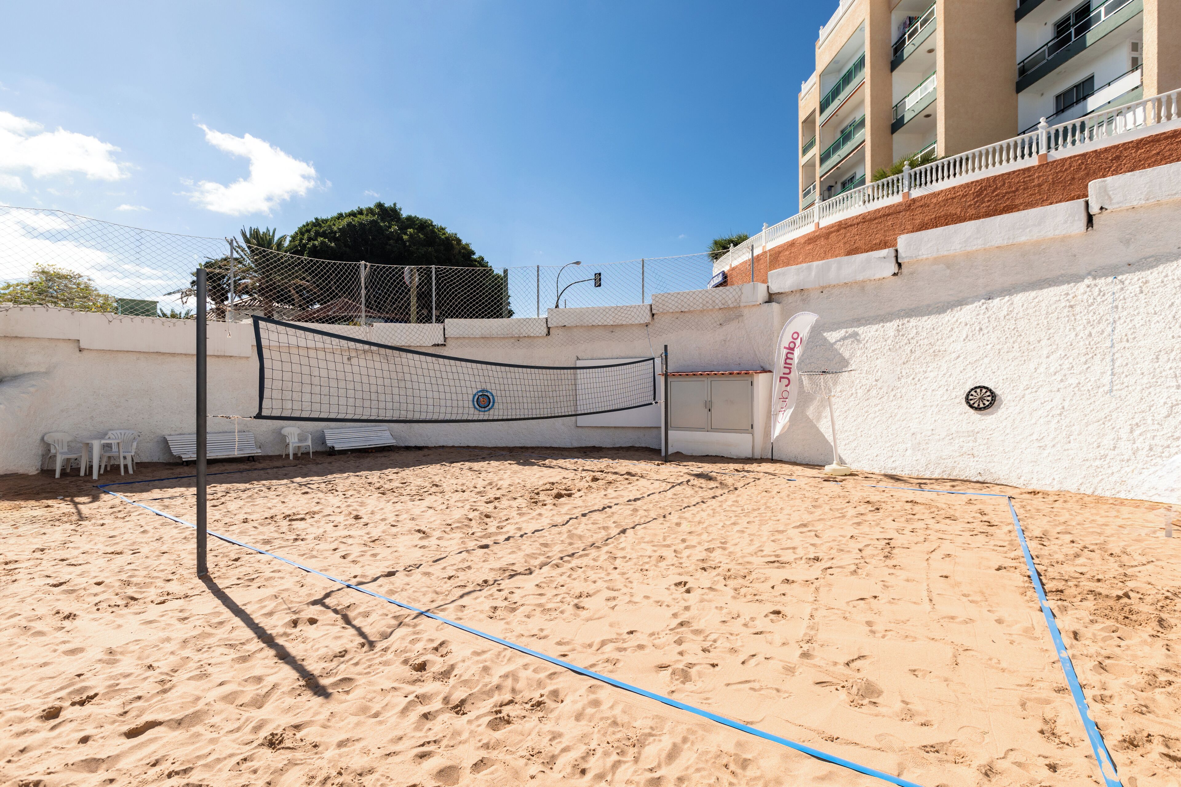 A beach volleyball court with a net, white chairs, and a building in the background.