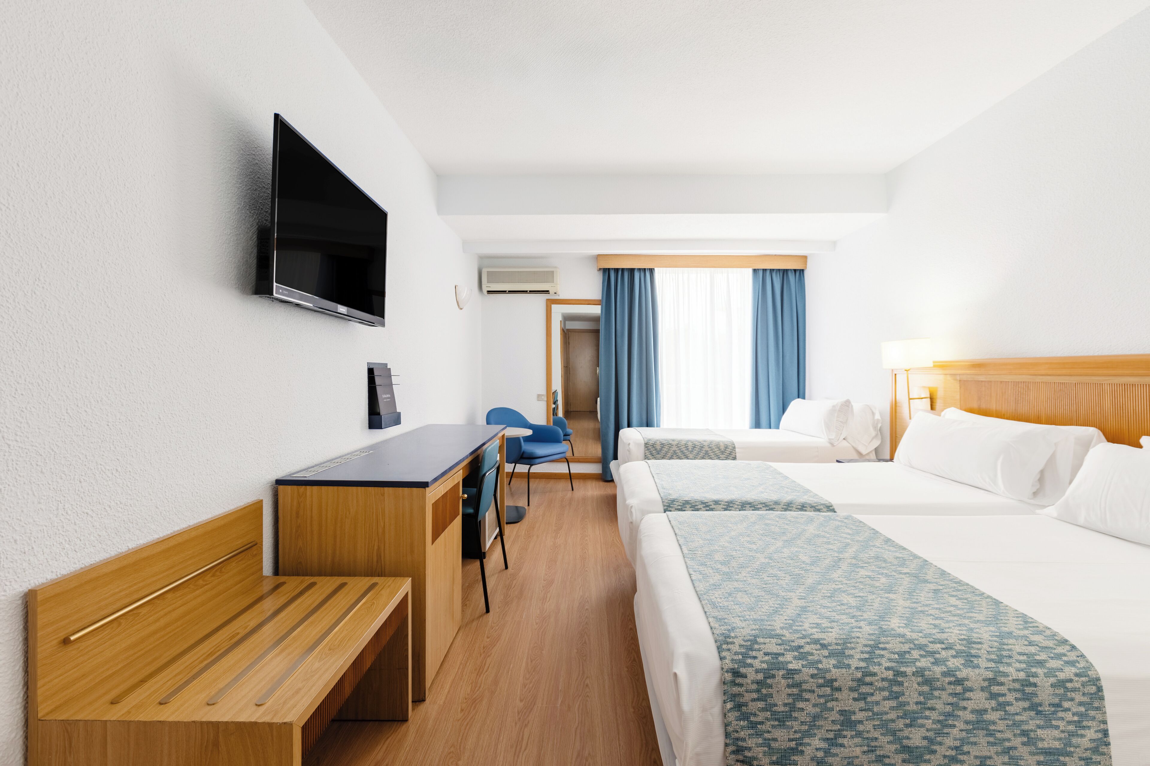 Een hotelkamer met een houten bureau, twee bedden, een televisie en een badkamer die door een deur zichtbaar is.