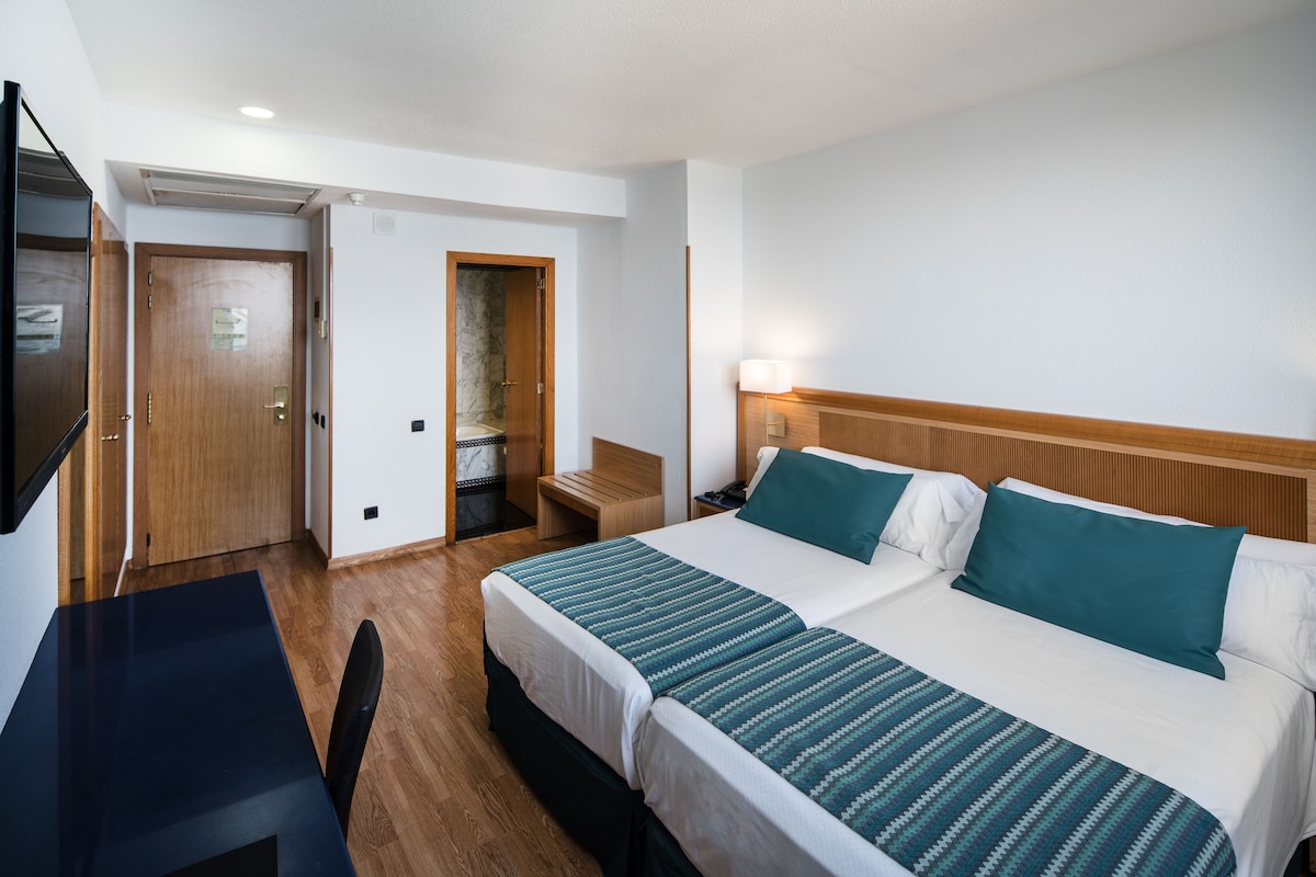 Hotellihuone, jossa on kaksi sänkyä, työpöytä, tuoli, puinen yöpöytä, televisio ja ovi, joka johtaa kylpyhuoneeseen.