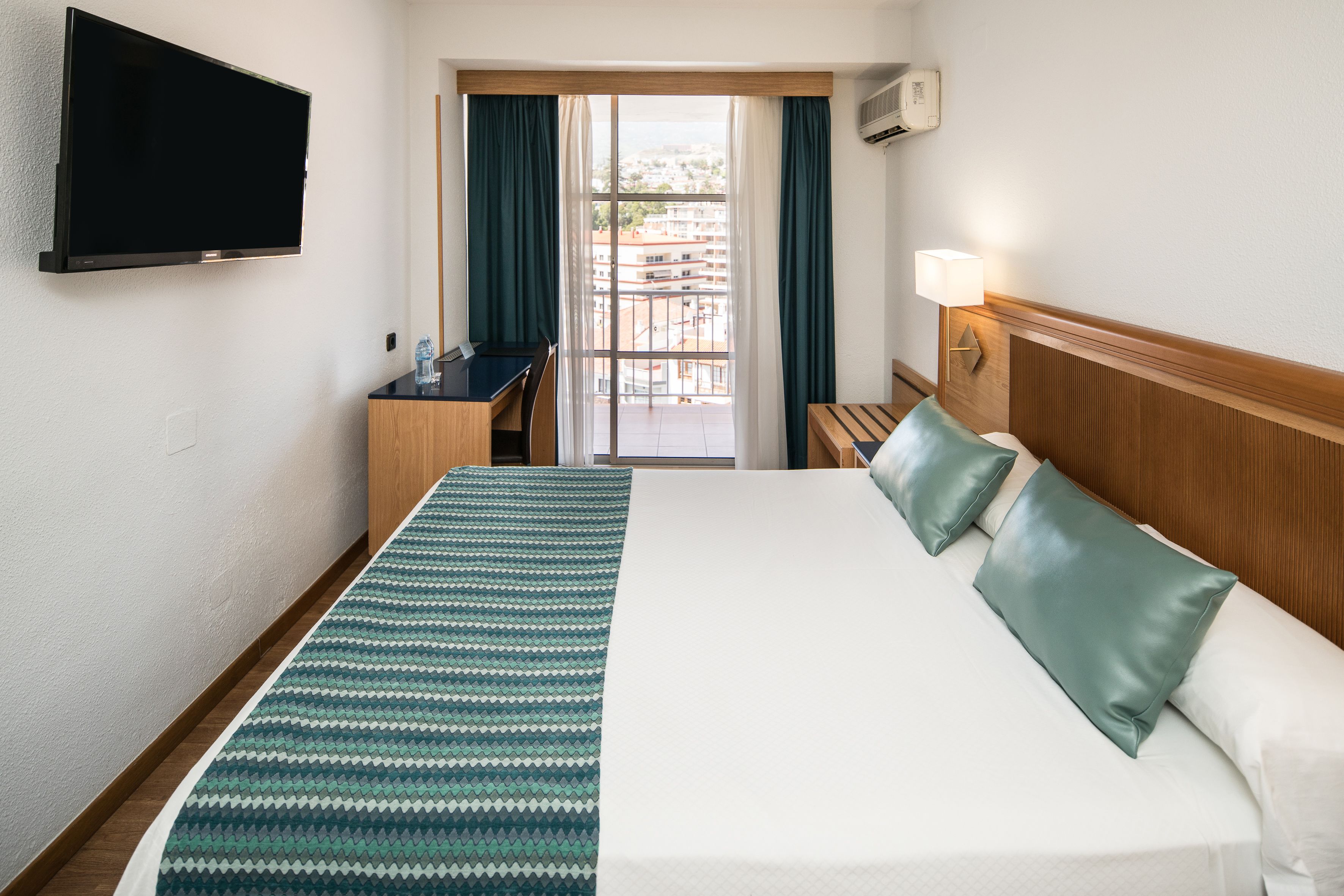 A hotel room with a bed, a desk, a chair, a TV, a window with curtains, and a view of buildings.