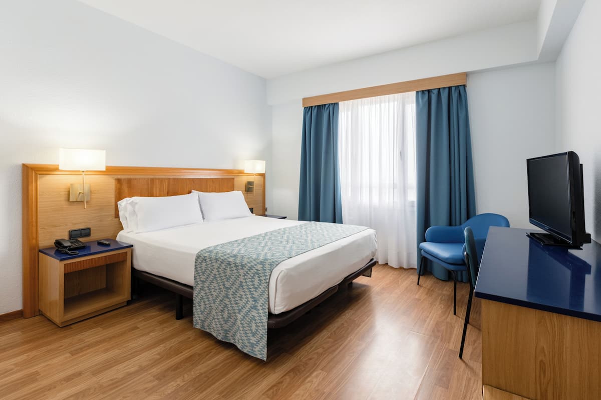 Una habitación de hotel con cama, mesitas de noche, un escritorio con televisor y una silla.