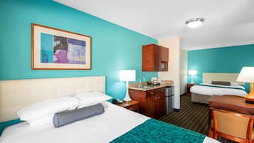 A hotel room with a teal wall, a bed with white bedding, a desk with a lamp, a kitchenette, and a framed picture on the wall.