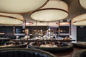 Um restaurante moderno com mesas redondas, assentos de couro preto e grandes luminárias pendentes.