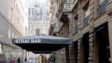 Straßenszene mit einem 'STRAF BAR'-Sonnenschirm, Außenbereich mit Sitzgelegenheiten und historischen Gebäuden im Hintergrund.