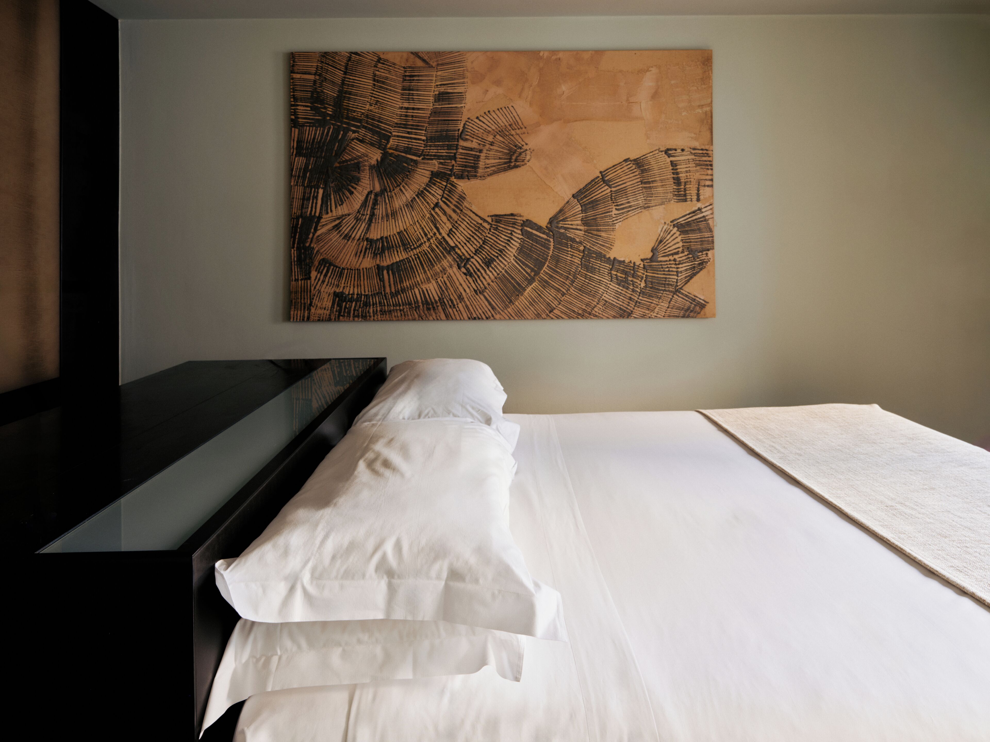 A modern hotel room with a large bed, a dark wooden dresser, and a large abstract painting on the wall.
