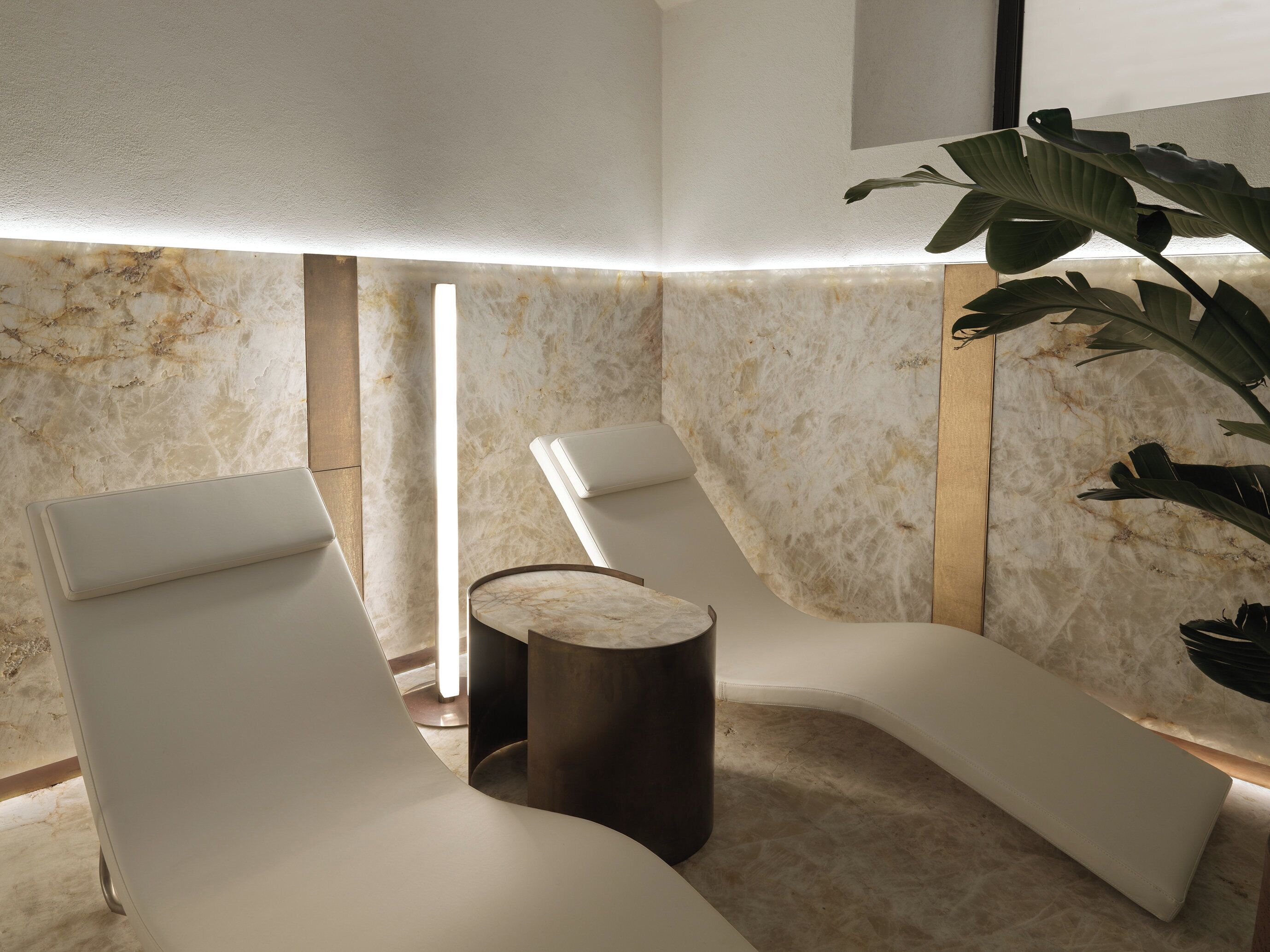 A modern spa area with marble walls, two white lounge chairs, a wooden side table, and a potted plant.