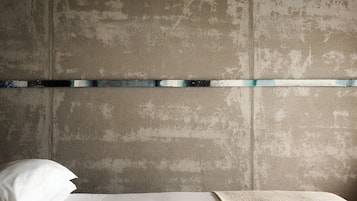 Un dormitorio con cabecera de concreto, una tira metálica y una colcha blanca.