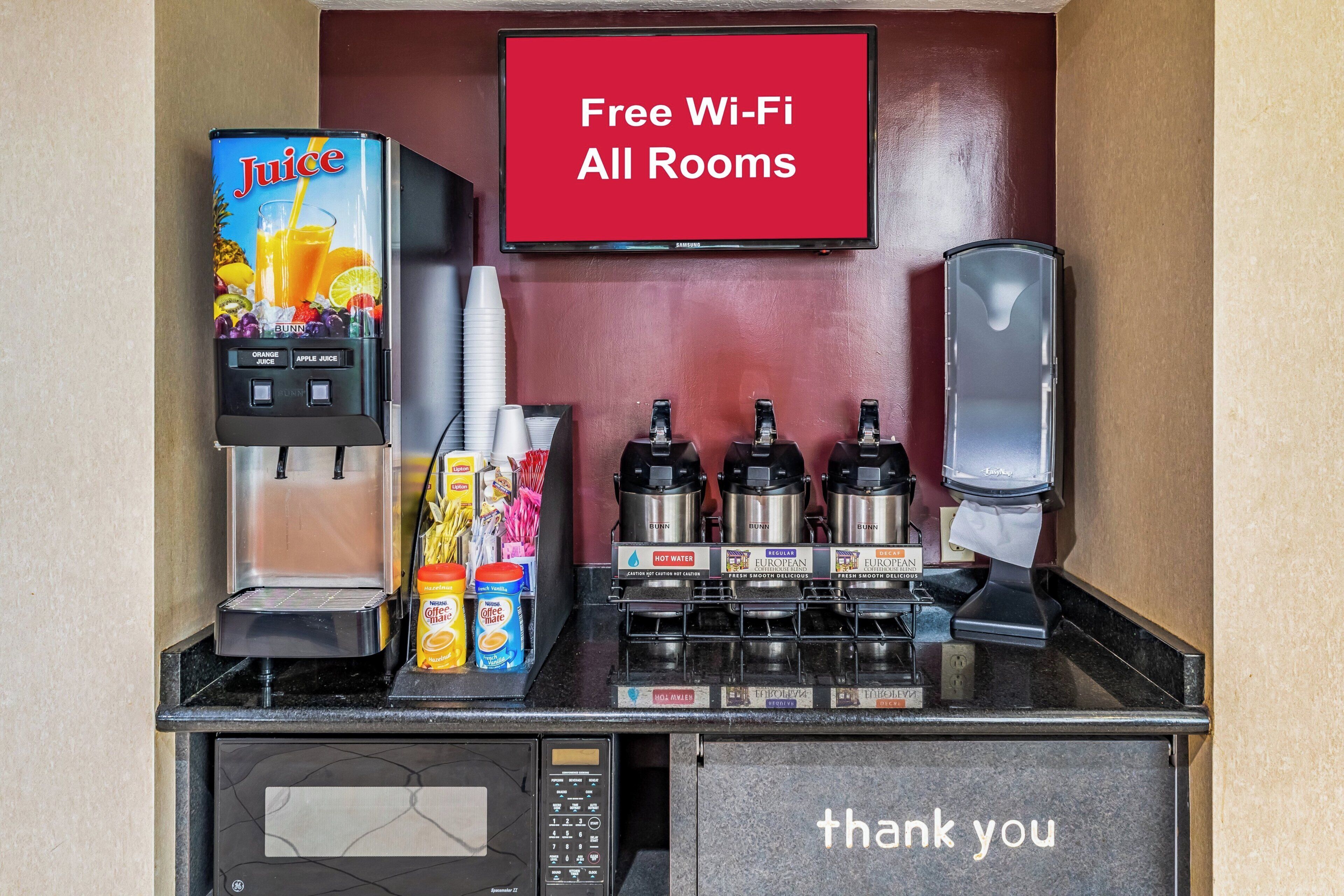 Eine Selbstbedienungs-Getränkestation mit Saftspender, Kaffeeautomaten und einer Mikrowelle. Ein Schild darüber zeigt, dass kostenloses WLAN in allen Zimmern verfügbar ist.