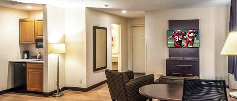 A hotel room with a kitchenette, a dining area with a table and chairs, a TV mounted on the wall, and a large mirror.