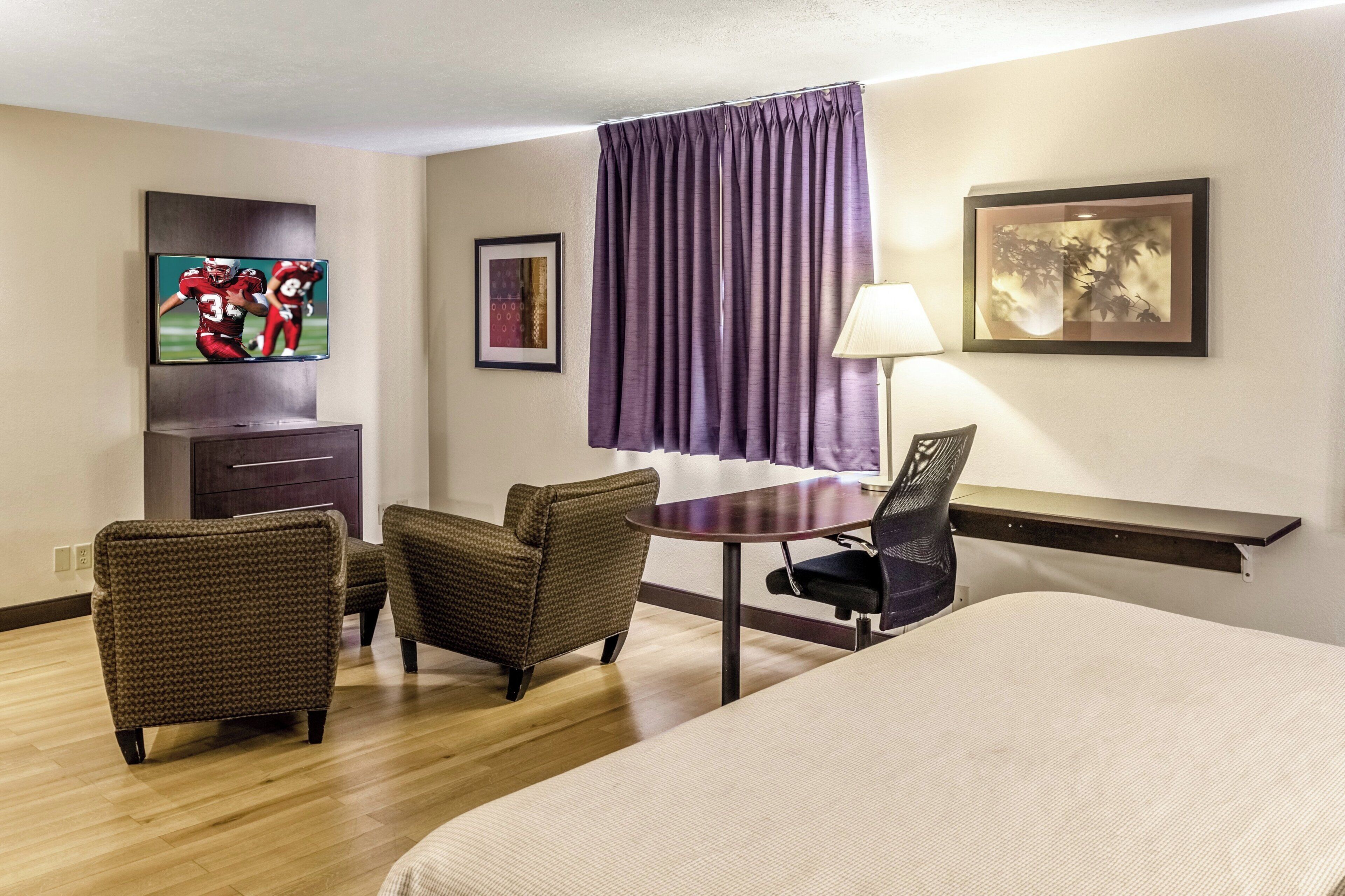 A hotel room with a bed, two armchairs, a desk with a lamp, a TV, and a window with curtains.