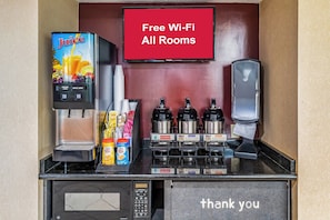 Una estación de bebidas de autoservicio con dispensador de zumo, agua caliente y cafeteras. Un cartel indica que hay Wi-Fi gratis en todas las habitaciones.