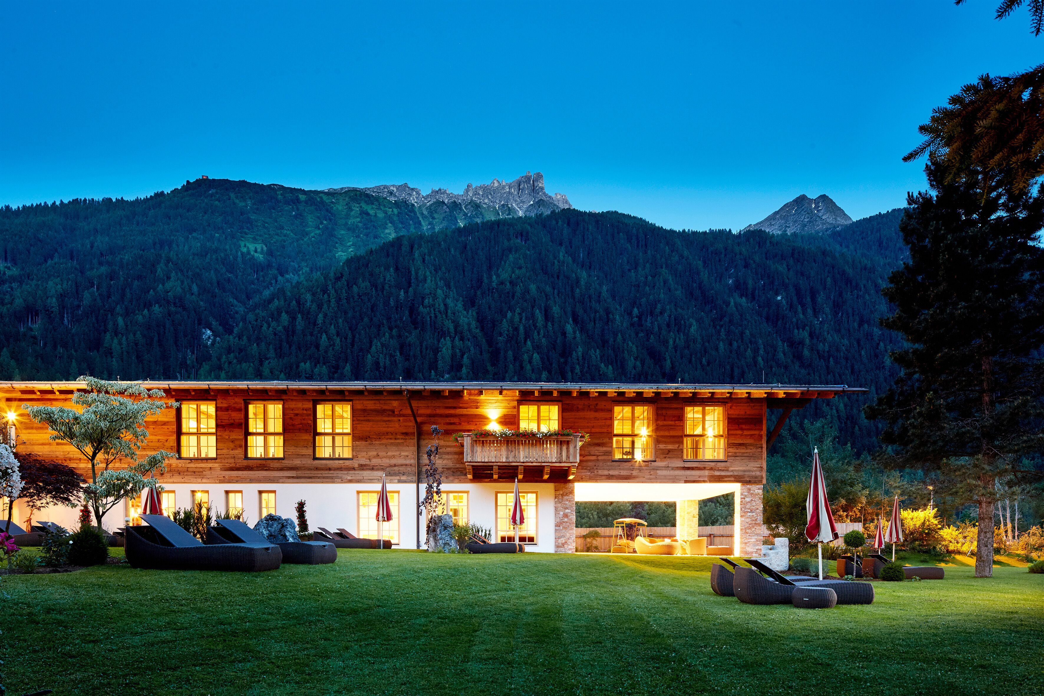 A modern wooden house with a large balcony, outdoor seating, and a mountain backdrop.