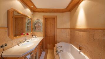 A bathroom with a double sink vanity, a large bathtub, wooden paneling, and a mirror with a decorative frame.