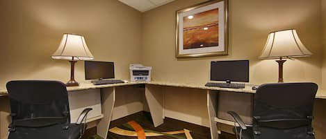 A hotel room with two desks, each with a computer, keyboard, and mouse. There are two chairs, a printer, and a framed picture on the wall.