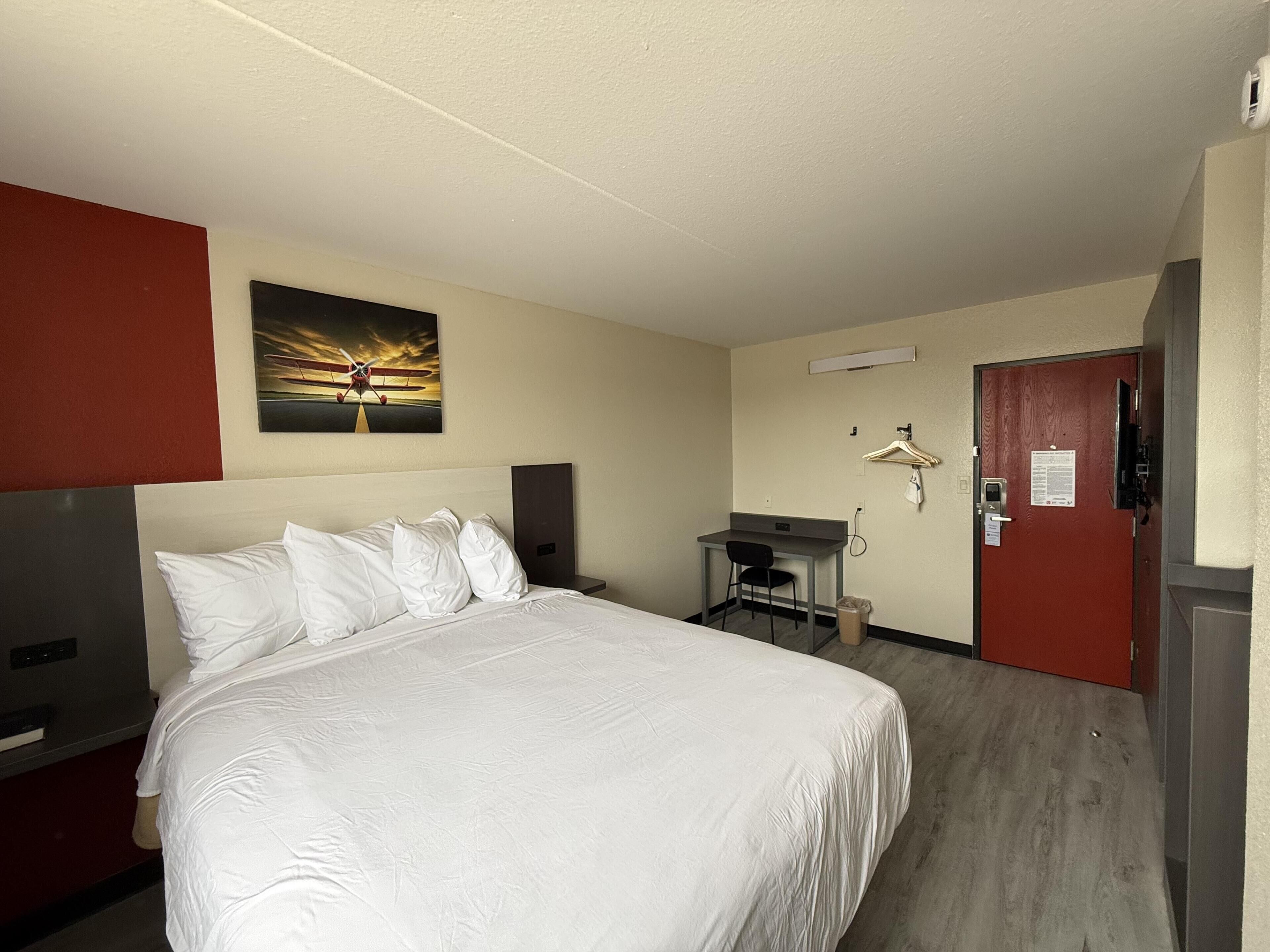 A hotel room with a bed, a desk, a chair, a coat rack, and a door.