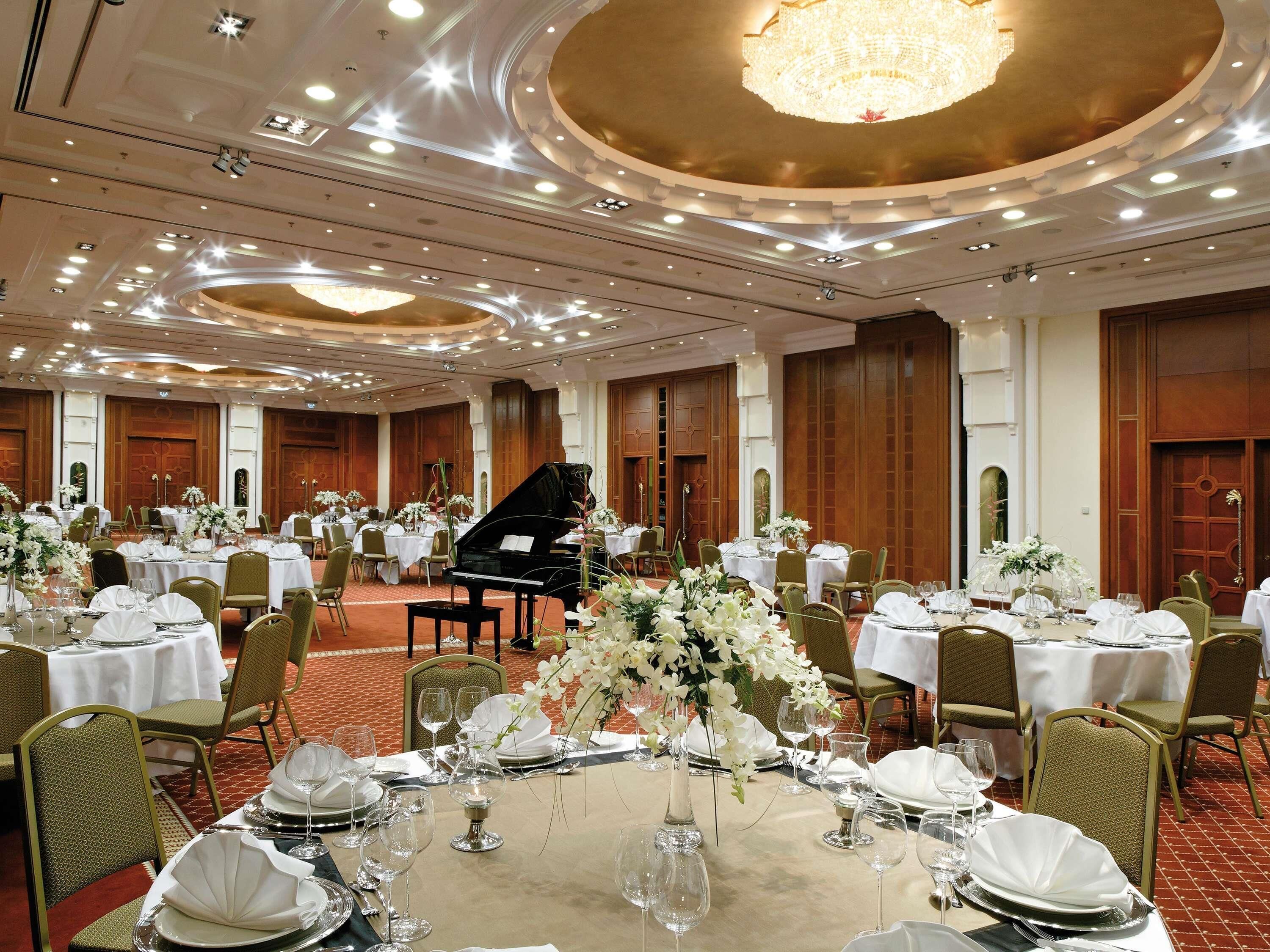 A banquet hall with round tables, chairs, a grand piano, and chandeliers.