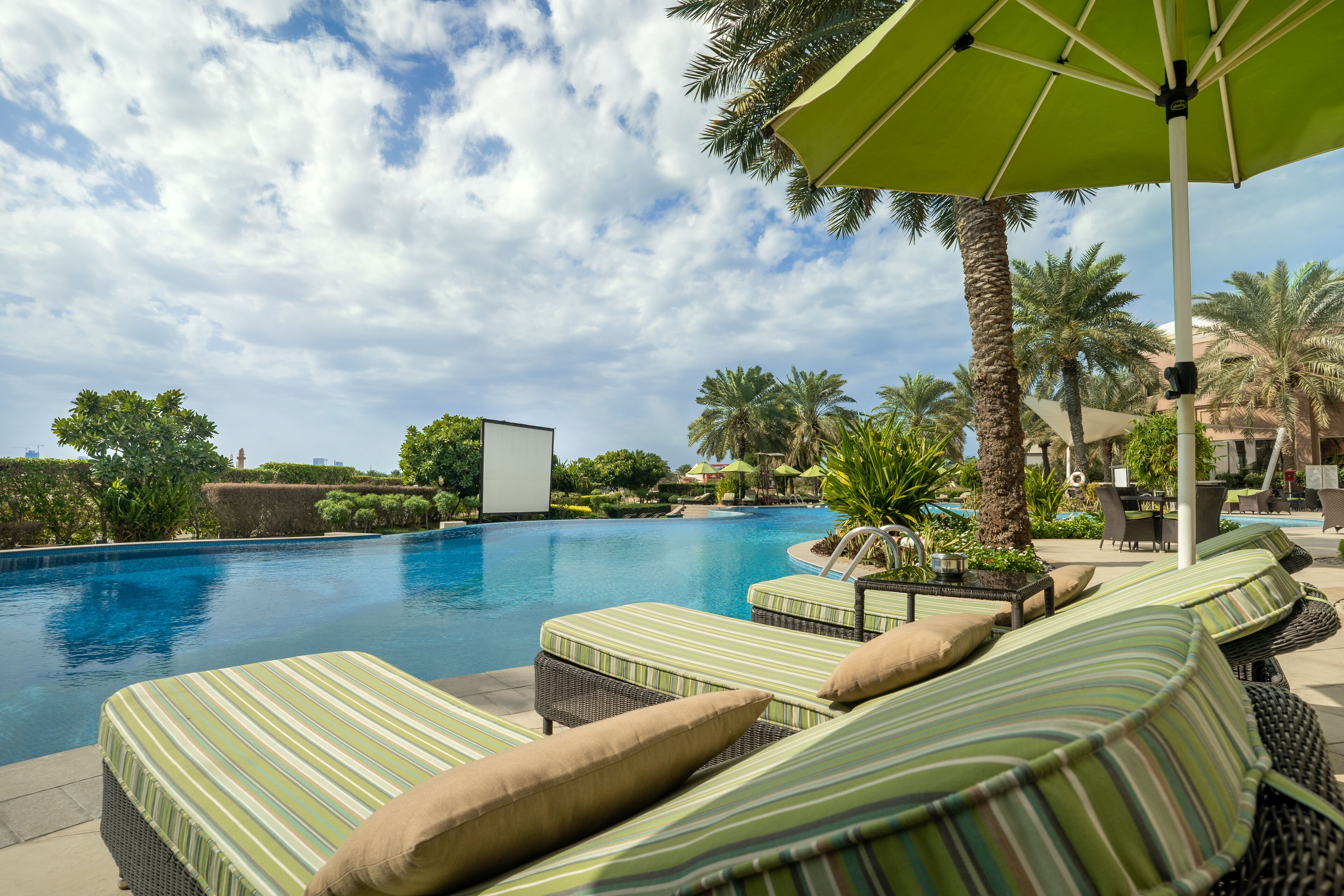 A poolside area with lounge chairs and umbrellas, surrounded by palm trees and a clear sky.
