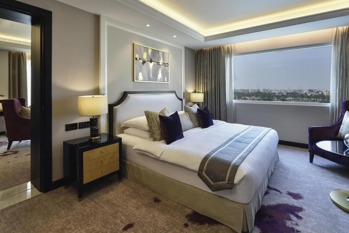 Habitación de hotel con una cama grande, mesitas de noche, un espejo y vistas de la ciudad, con un paisaje urbano visible a través de la ventana.