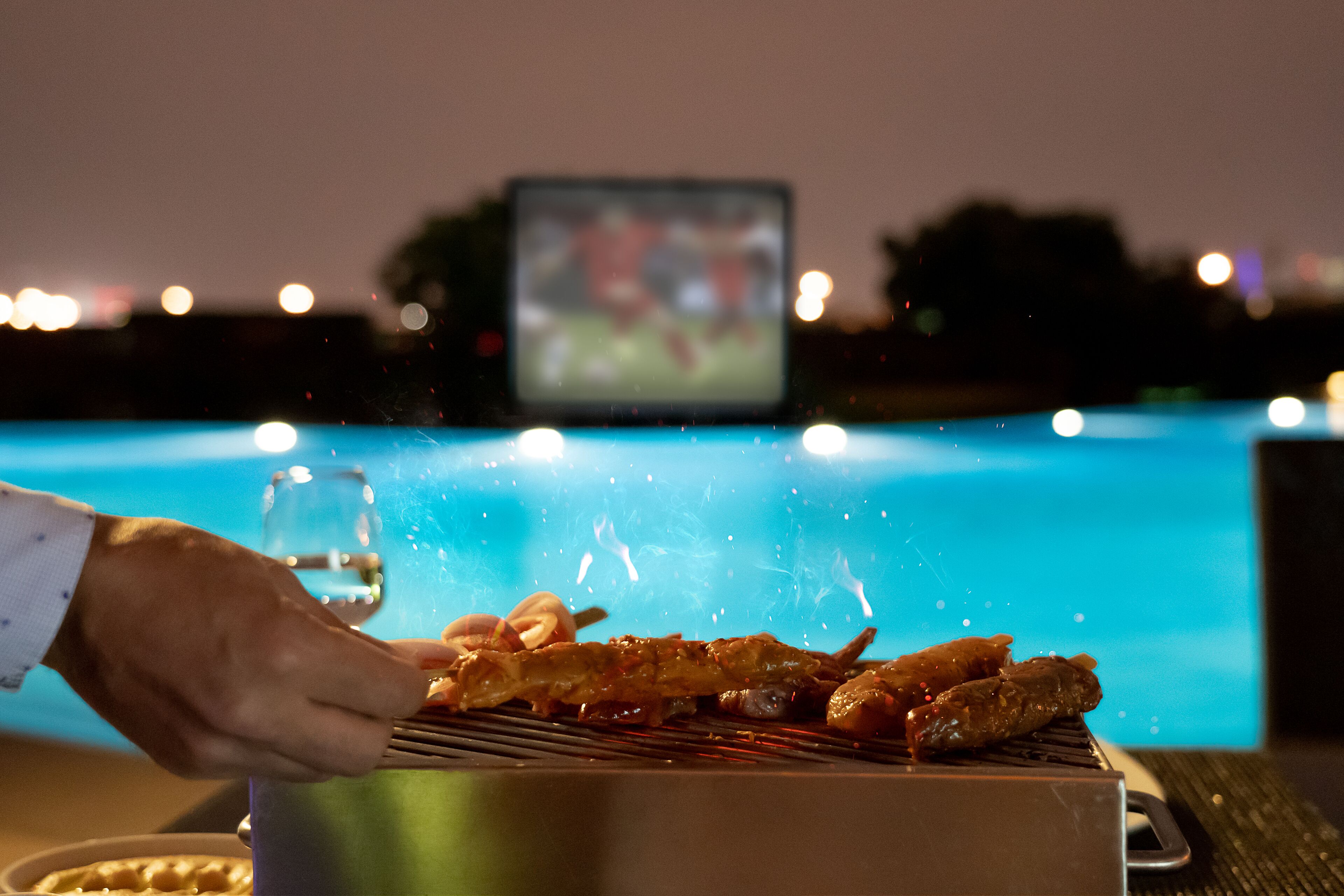 Une personne qui prépare des aliments sur un barbecue au bord d’une piscine, avec un téléviseur en arrière-plan.