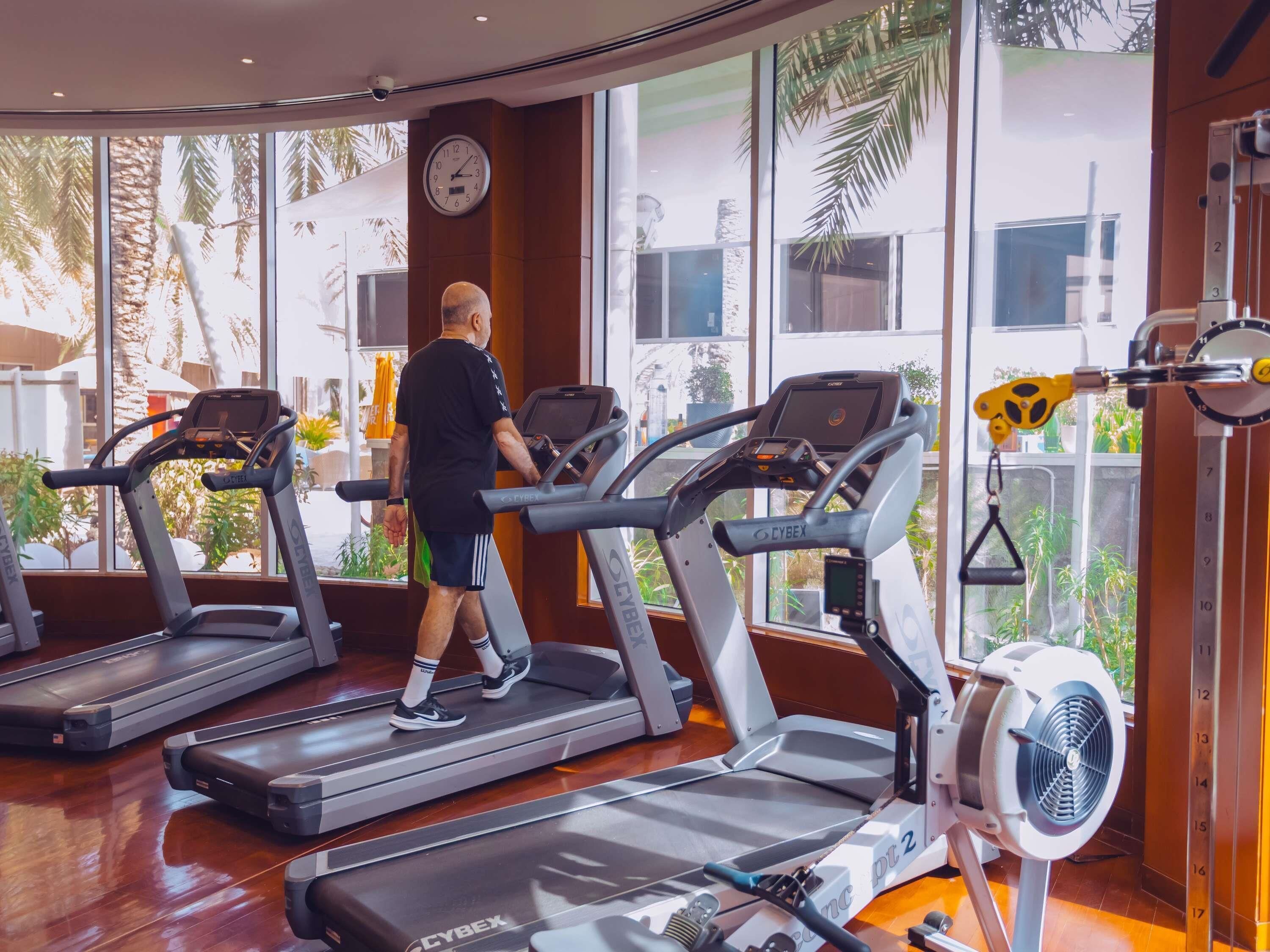 A gym with treadmills, a wall clock, and large windows with a view of palm trees.